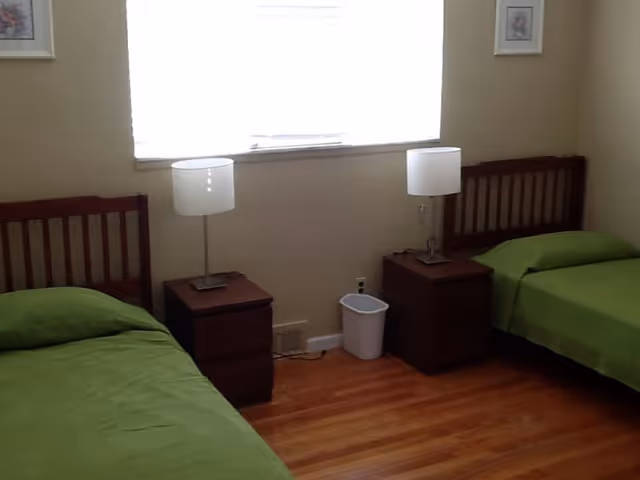 A simple bedroom with two single beds, each with a green bedspread. Between the beds are two wooden nightstands, each with a white lamp. There is a window with blinds above the nightstands, and the floor is wooden.