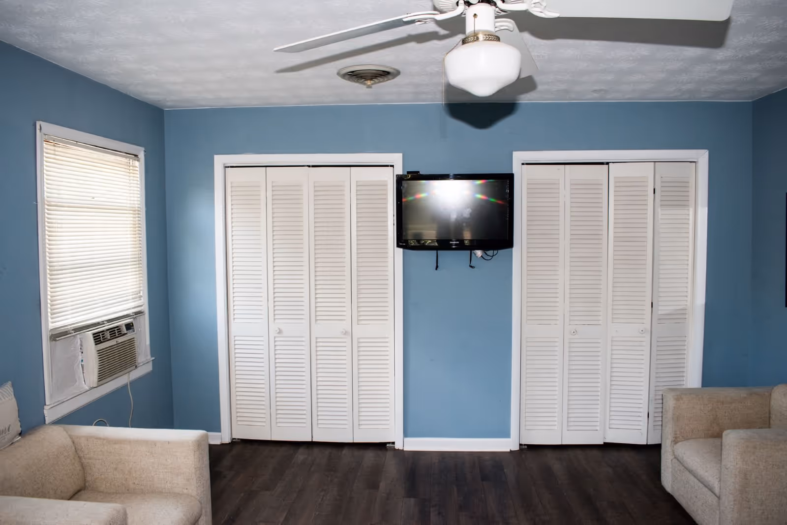 A living room with blue walls, two beige armchairs, a window with blinds and an air conditioning unit, two white louvered closet doors, a wall-mounted flat-screen TV, and a ceiling fan with a light fixture.