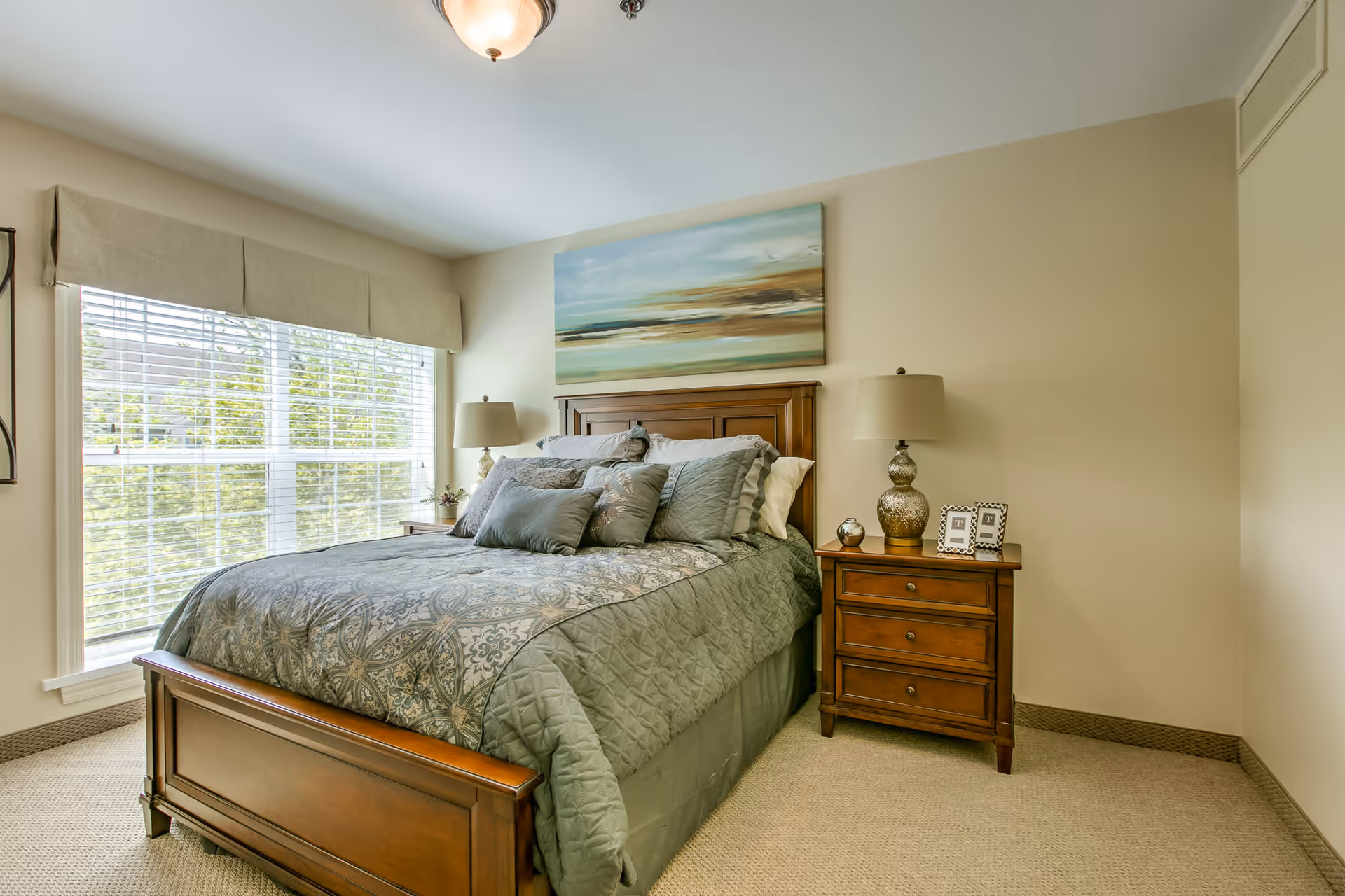 A bedroom with a large wooden bed featuring a patterned gray and blue comforter and multiple pillows. There are two matching wooden nightstands on either side of the bed, each with a lamp. A large window with white blinds and a beige valance lets in natural light. Above the bed hangs a landscape painting with soft blue, green, and brown tones. The walls are painted a light beige color, and the floor is carpeted.