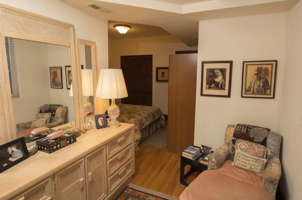 A cozy bedroom area with a floral armchair and a matching ottoman, a small side table with books and a photo frame, a dresser with a large mirror, a table lamp, and framed pictures on the walls. The bed with a floral bedspread is partially visible in the background.