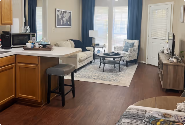 Open-plan living room and kitchenette with a sofa, armchair, coffee table, TV console, and breakfast bar in front of large windows and a door.