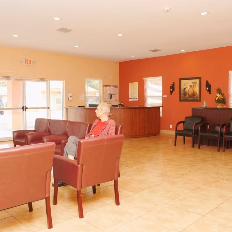 A senior man sitting on a maroon armchair in a spacious waiting area with tiled floors, maroon leather chairs, a wooden reception desk, and walls painted in beige and orange. There are windows with blinds, a framed painting, and some chairs lined up against the wall.