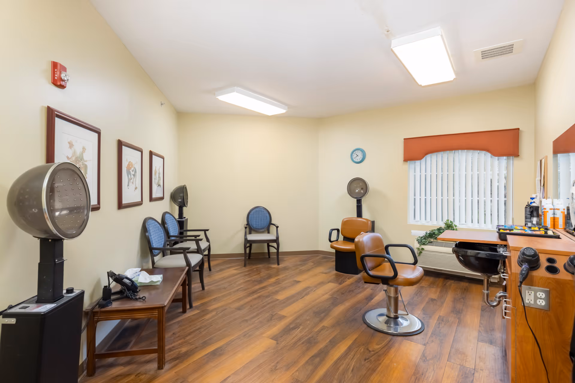 A hair salon room with wooden flooring, two brown salon chairs, two hair dryers, a counter with hair products and a hair dryer, three blue chairs along the wall, framed pictures on the wall, a window with vertical blinds and a red valance, and a clock on the wall.