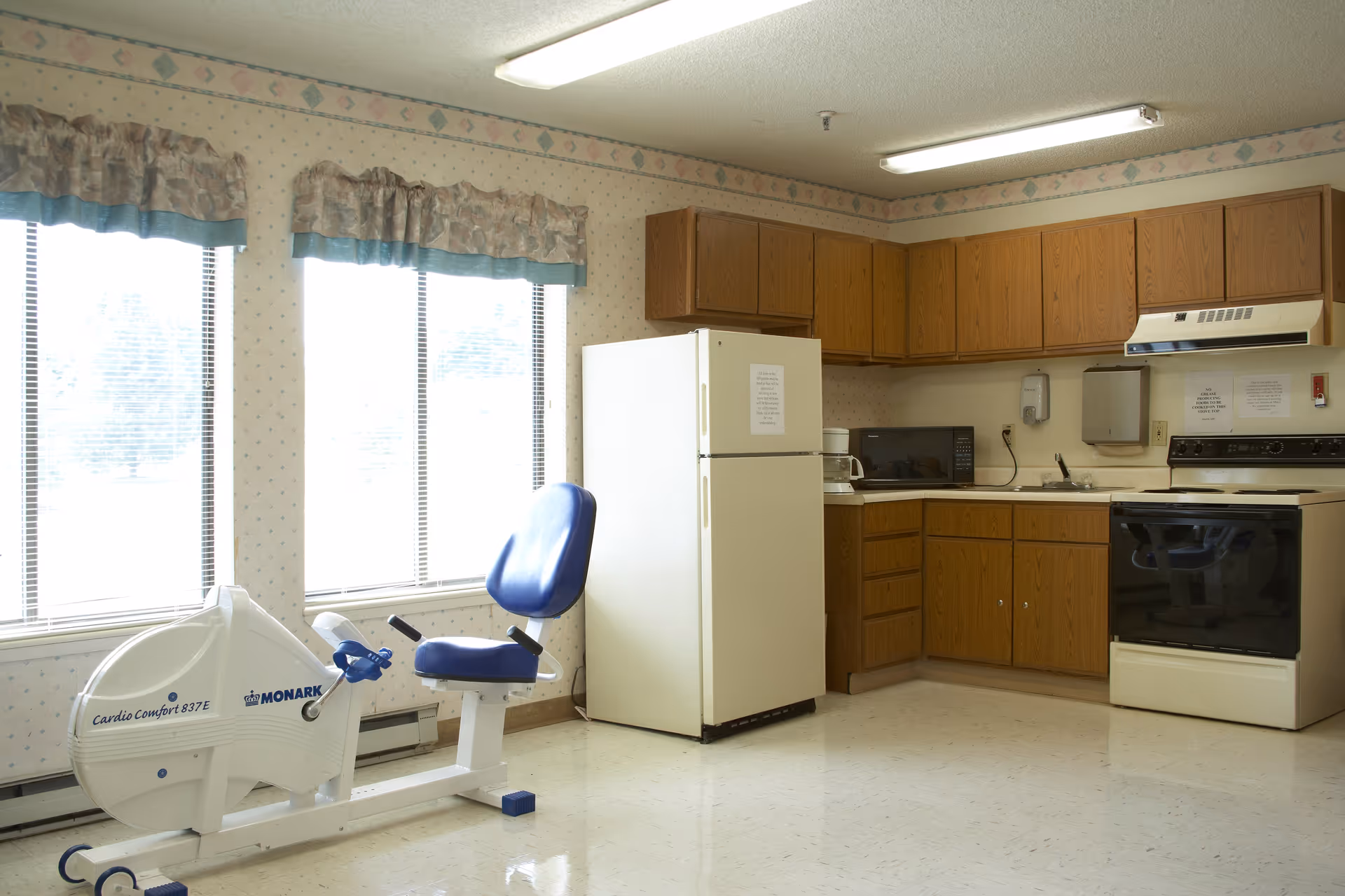 A kitchen area with wooden cabinets, a white refrigerator, a microwave, a stove with an overhead vent, and a white exercise bike with a blue seat positioned near three large windows with floral valances.