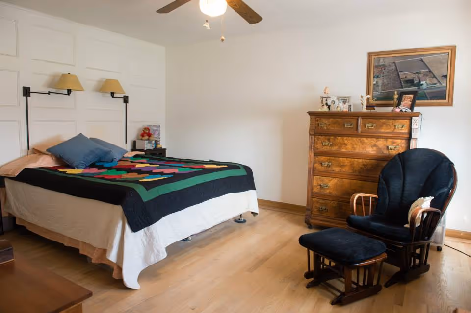 A tidy bedroom with a bed topped by a colorful patchwork quilt, a wooden dresser, and a cushioned rocking chair with ottoman on hardwood floors.
