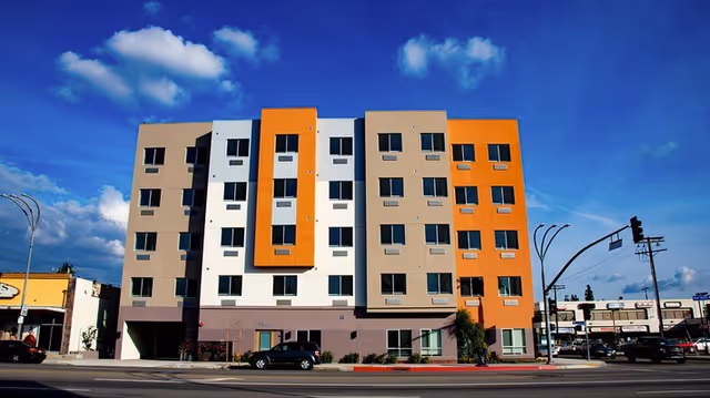 Exterior view of a modern multi-story senior living facility building with beige, white, and orange panels under a blue sky with scattered clouds. There are cars parked in front and a traffic light on the right side of the image.