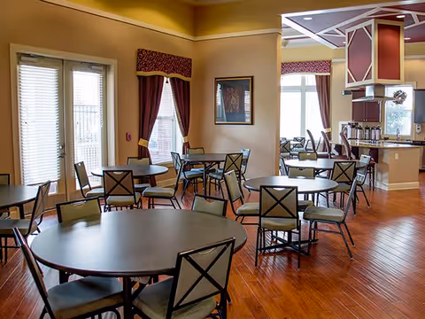 A spacious dining area with multiple round tables and chairs arranged neatly on a polished wooden floor. Large windows with blinds and burgundy curtains allow natural light to fill the room. The walls are painted in warm tones, and there is a framed picture on one wall. The room also features a kitchen area with a counter and coffee dispensers visible in the background.