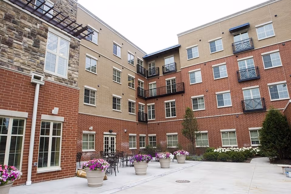 Outdoor courtyard area of Lombard Place Assisted Living & Memory Care with potted flowers, patio tables and chairs, surrounded by a multi-story brick and stone building with multiple windows and small balconies.