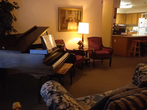 Cozy living room area with a grand piano, two upholstered chairs, a side table with a lamp, and a floral patterned sofa. In the background, there is a kitchen with wooden cabinets, a refrigerator, and a counter with bar stools.