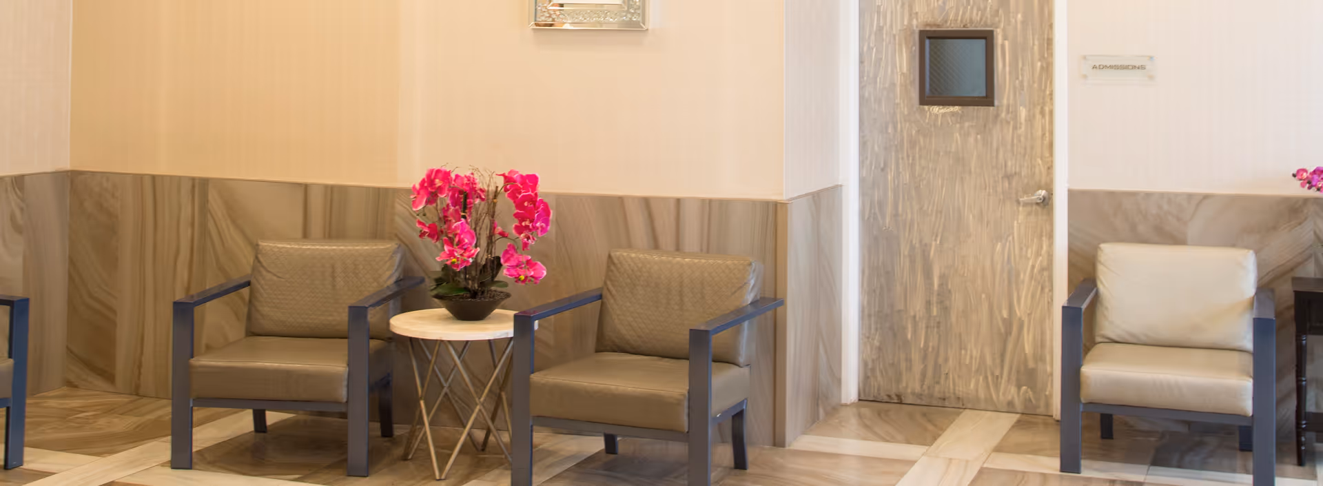 A waiting area with three cushioned chairs arranged around a small round table holding a vibrant pink orchid plant. The walls have a beige and marble pattern, and there is a door labeled 'Admissions' in the background.