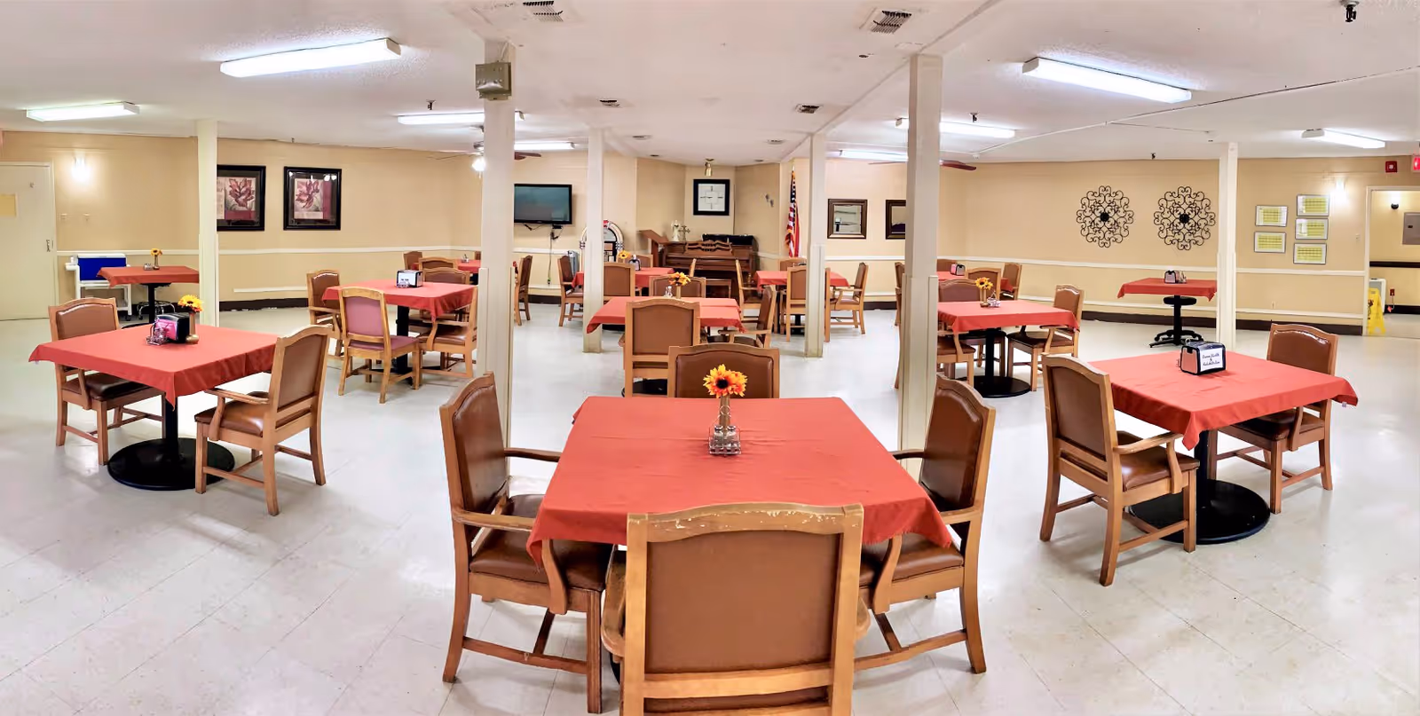 A spacious dining room with multiple tables covered in red tablecloths, each surrounded by wooden chairs with brown cushions. The room has beige walls decorated with framed pictures and wall art, a television mounted on the wall, and an American flag in the background. The floor is light-colored tile, and the ceiling has fluorescent lighting.