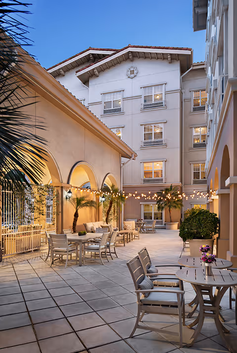Outdoor courtyard area of Belmont Village Senior Living Burbank with tiled flooring, several tables and chairs, potted plants, string lights hanging overhead, and a multi-story building in the background during dusk.