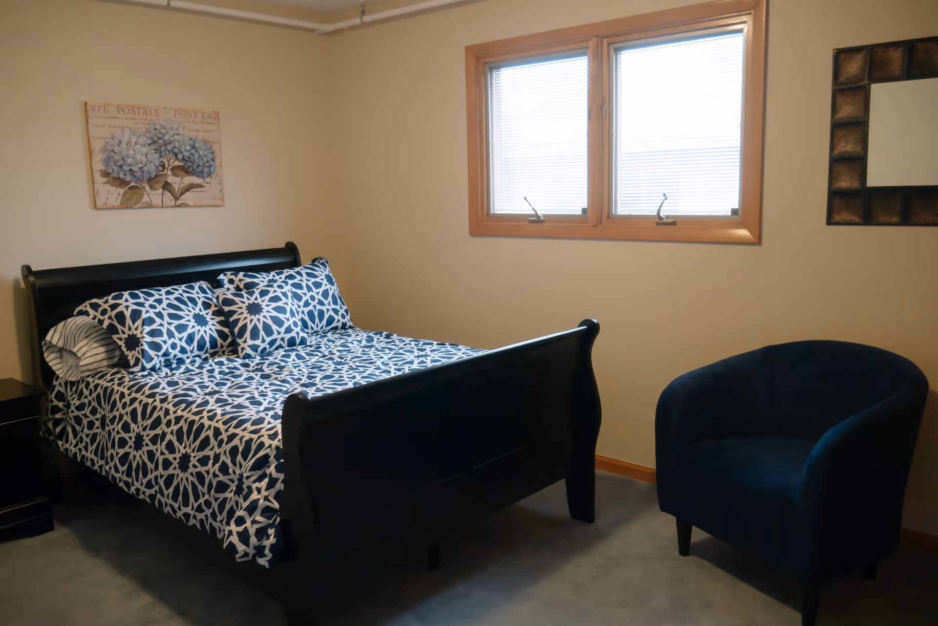 A bedroom with a black wooden bed frame featuring a blue and white patterned bedspread and pillows. There is a dark blue upholstered armchair to the right, a window with blinds above the chair, a framed floral artwork on the wall above the bed, and a decorative mirror on the wall to the right.