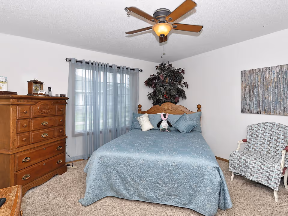 A cozy bedroom with a wooden bed frame and a blue quilted bedspread, decorated with pillows and a stuffed animal. There is a wooden dresser with drawers on the left side, a window with sheer blue curtains behind the bed, a ceiling fan with a light fixture above, a potted plant behind the bed, and a patterned armchair on the right side. A piece of artwork hangs on the wall above the armchair.
