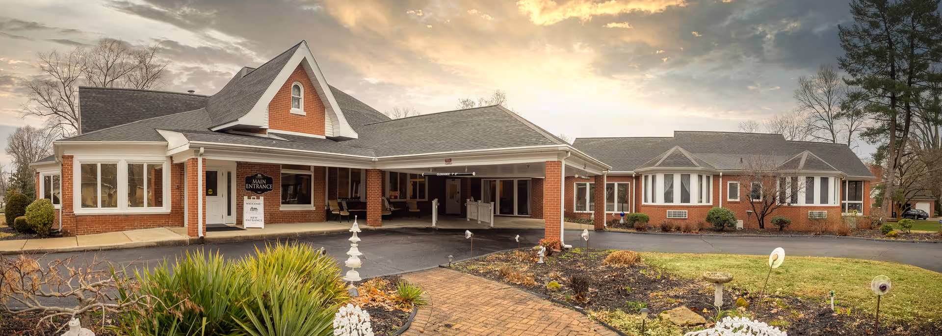 Exterior view of a brick building with a covered entrance labeled 'Main Entrance' at Charles Ford Retirement Communities of New Harmony. The building is surrounded by landscaped gardens and a paved driveway under a cloudy sky during sunset.