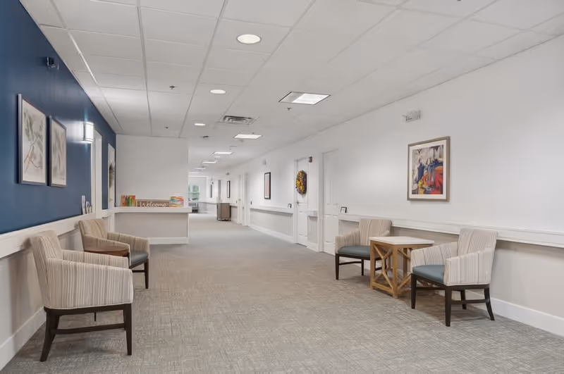 A long, well-lit hallway in a senior living facility with beige carpet and white walls. The left wall is painted dark blue and decorated with framed artwork. There are several pairs of upholstered chairs and small wooden tables arranged along the hallway. Doors and additional framed pictures line the right wall, and a welcome sign is visible on a counter at the far end.