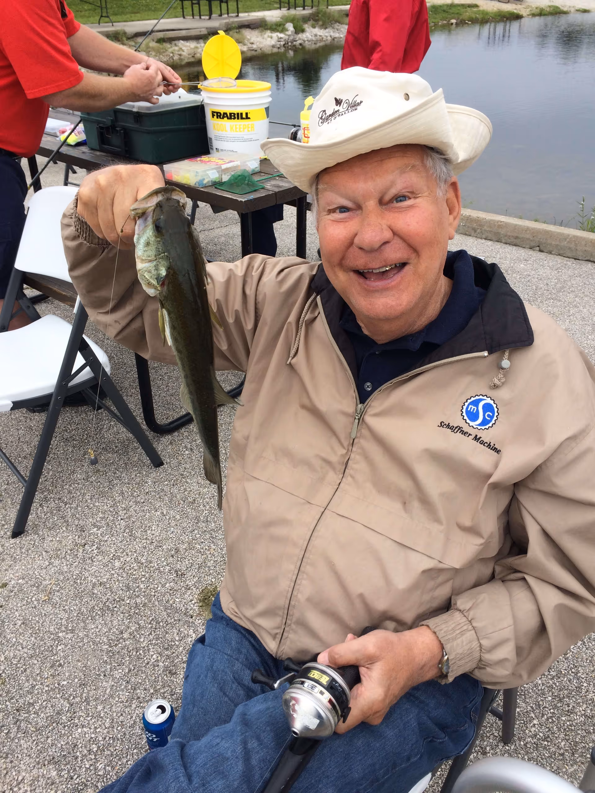 An elderly man wearing a beige jacket and a white hat with the Garden Villas of O’Fallon logo is sitting outdoors near a pond, smiling and holding up a fish he caught with a fishing rod.