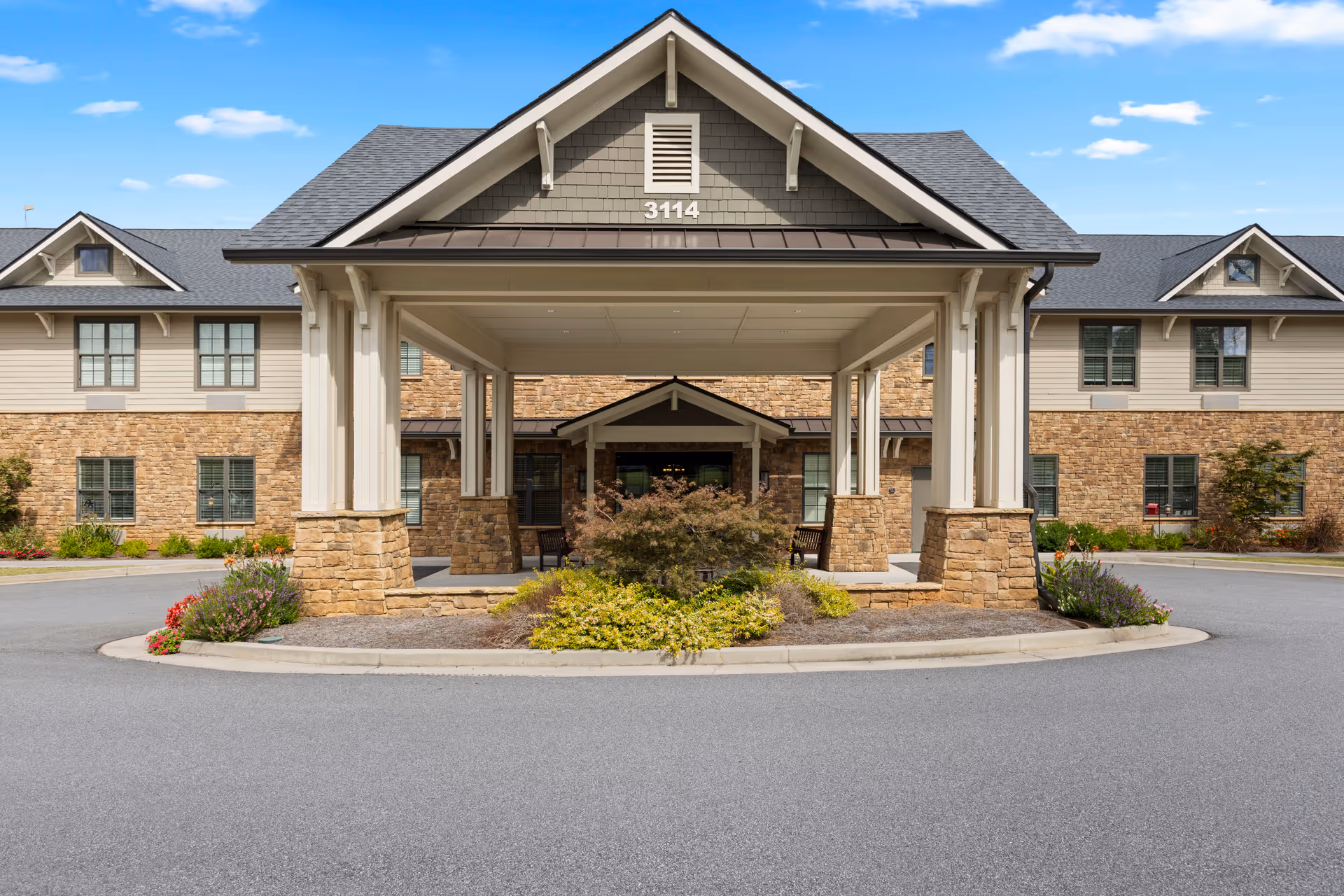 Front entrance of a senior living facility with a covered drop-off area supported by stone and white pillars, surrounded by landscaped plants and flowers, under a blue sky with some clouds.