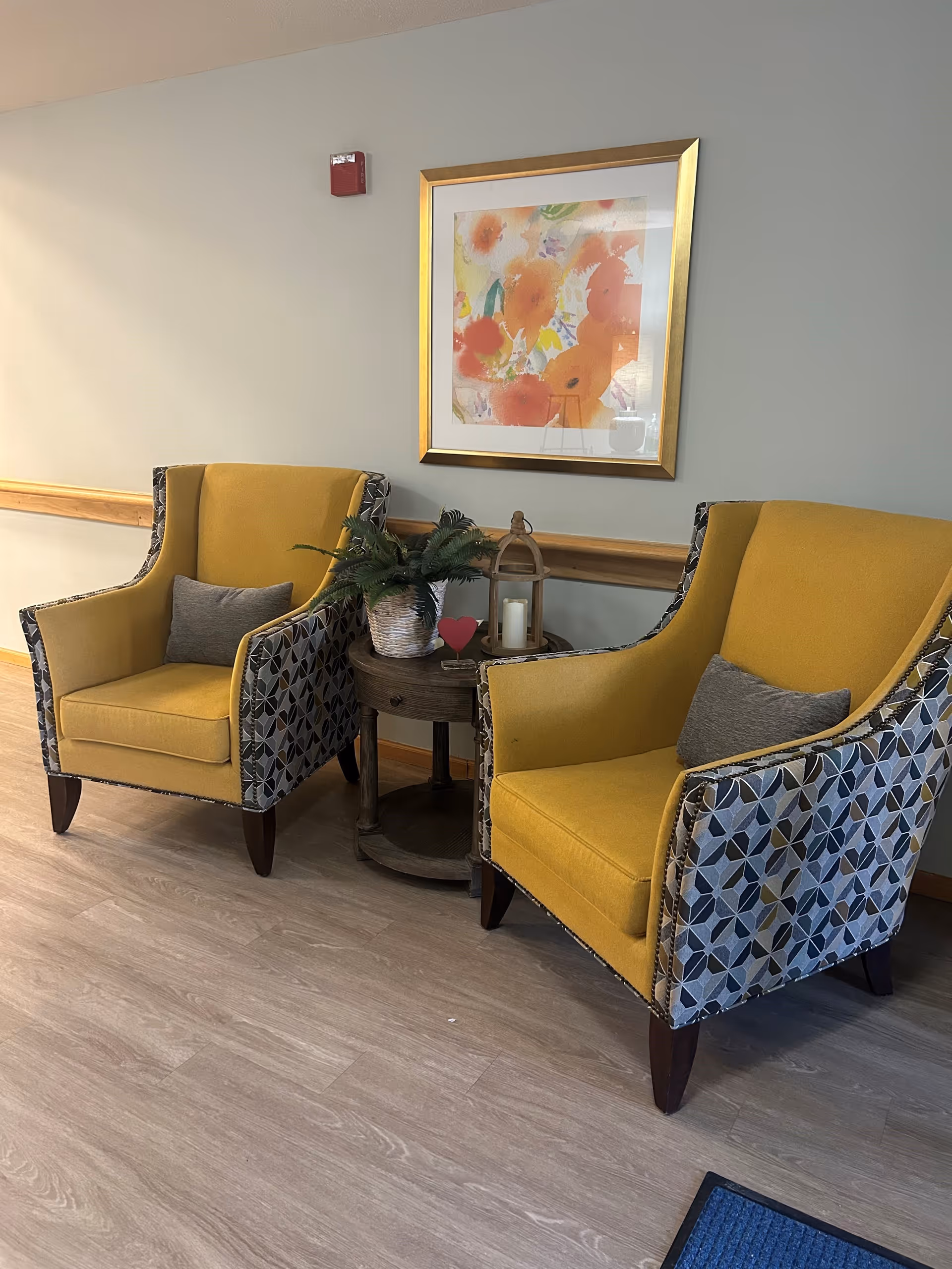 Two yellow armchairs with patterned sides and gray pillows are placed on either side of a small round wooden table. The table holds a potted plant and a decorative lantern with a candle. Above the chairs, a framed abstract floral painting with orange and yellow hues hangs on a light gray wall. The floor is light wood, and a wooden chair rail runs along the wall behind the chairs.