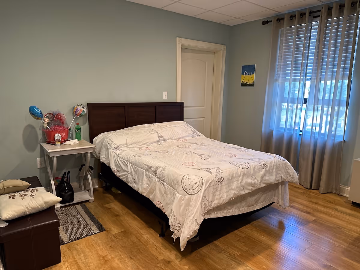 Neat bedroom with a double bed and wooden headboard, a nightstand with gifts and balloons, and a curtained window letting in daylight.