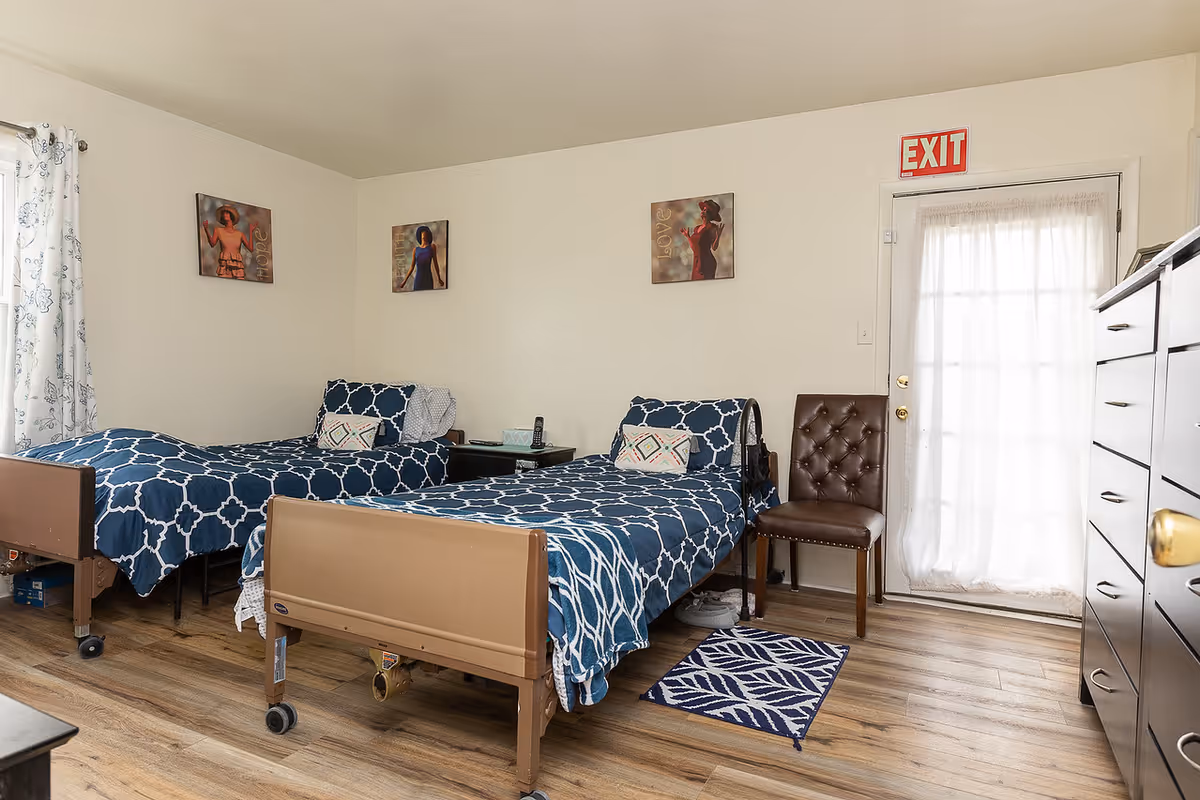 A bedroom in a senior living facility with two single beds covered in blue and white patterned bedding. There is a brown chair between the beds and a dresser on the right side. The room has wooden flooring, three framed pictures on the wall, a window with white curtains on the left, and a door with a sheer curtain and an exit sign above it.