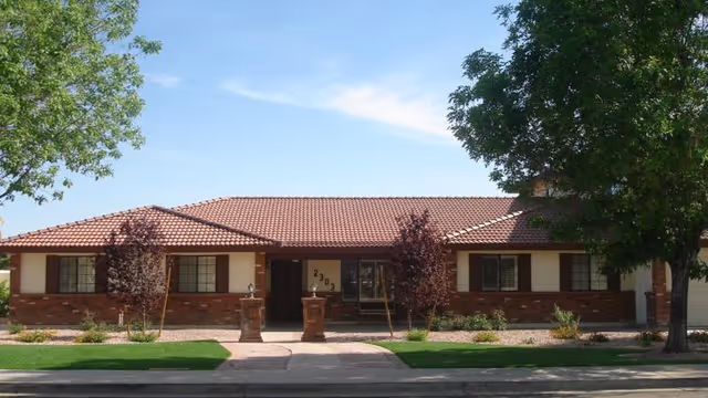 Single-story residential building with a red tile roof, brick and beige exterior walls, and several windows with brown shutters. The front entrance has two brick pillars and the number 2303 displayed above the door. There are trees and landscaping with grass and small bushes in front of the building.