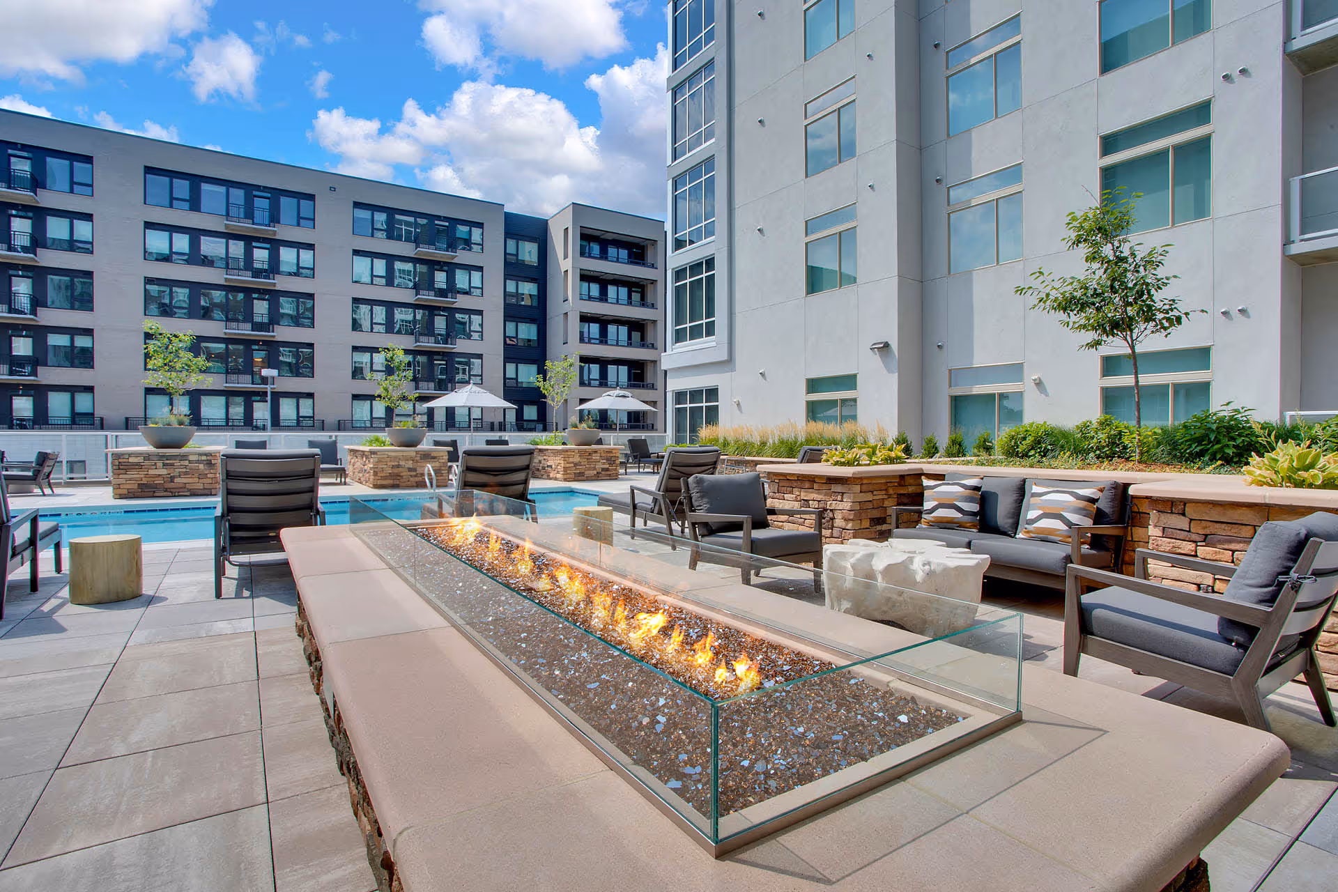 Outdoor patio area at Overture 9th + Co 55+ Apartment Homes featuring a modern rectangular fire pit with glass wind guard, surrounded by cushioned seating and chairs. In the background, there is a swimming pool with lounge chairs and umbrellas, and multi-story apartment buildings under a partly cloudy blue sky.
