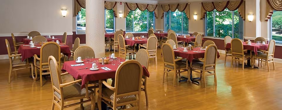 A spacious dining room with multiple tables covered in red tablecloths, each set with white cups, saucers, and silverware. The room has wooden floors, large windows with decorative curtains, and wall-mounted lights providing warm illumination.
