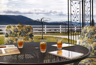 Outdoor patio area with a round metal table holding four glasses of iced tea and some books or magazines. The table is surrounded by cushioned chairs with floral patterned upholstery. In the background, there is a white fence, some bare trees, and a scenic view of mountains and a cloudy sky.