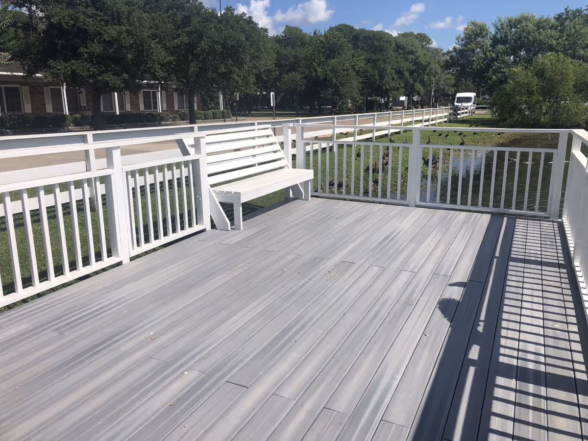 A wooden deck with white railings and a white bench overlooking a small pond with ducks. Trees and a building are visible in the background under a blue sky with some clouds.