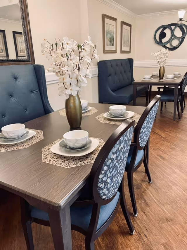 A dining room with two wooden tables set with white and gray marbled bowls and plates on beige placemats. Each table has a gold vase with white flowers as a centerpiece. The chairs have blue upholstery with a patterned backrest, and there are blue tufted bench seats against the wall. The room has wooden flooring, framed artwork on the walls, and a decorative wall piece.