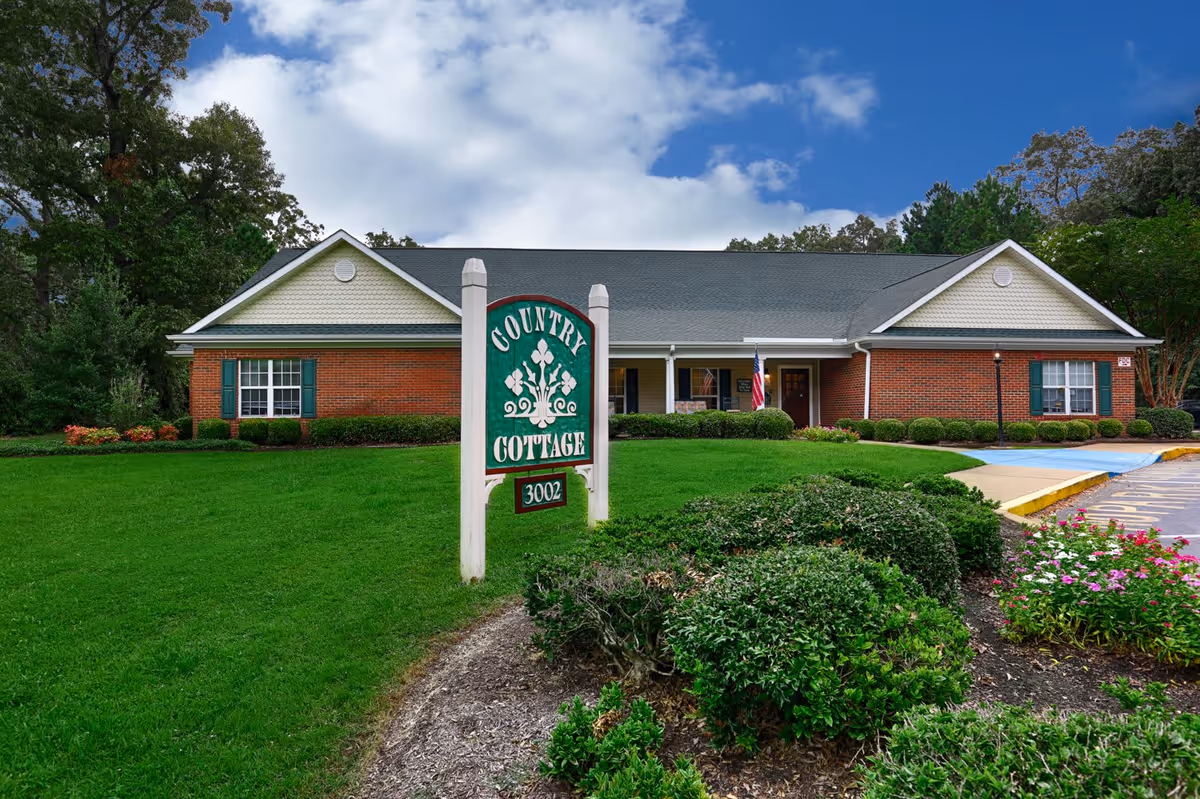 Exterior view of a single-story brick building with a gray roof, surrounded by green grass, bushes, and flowers under a partly cloudy sky. A white sign in front reads 'Country Cottage 3002'.