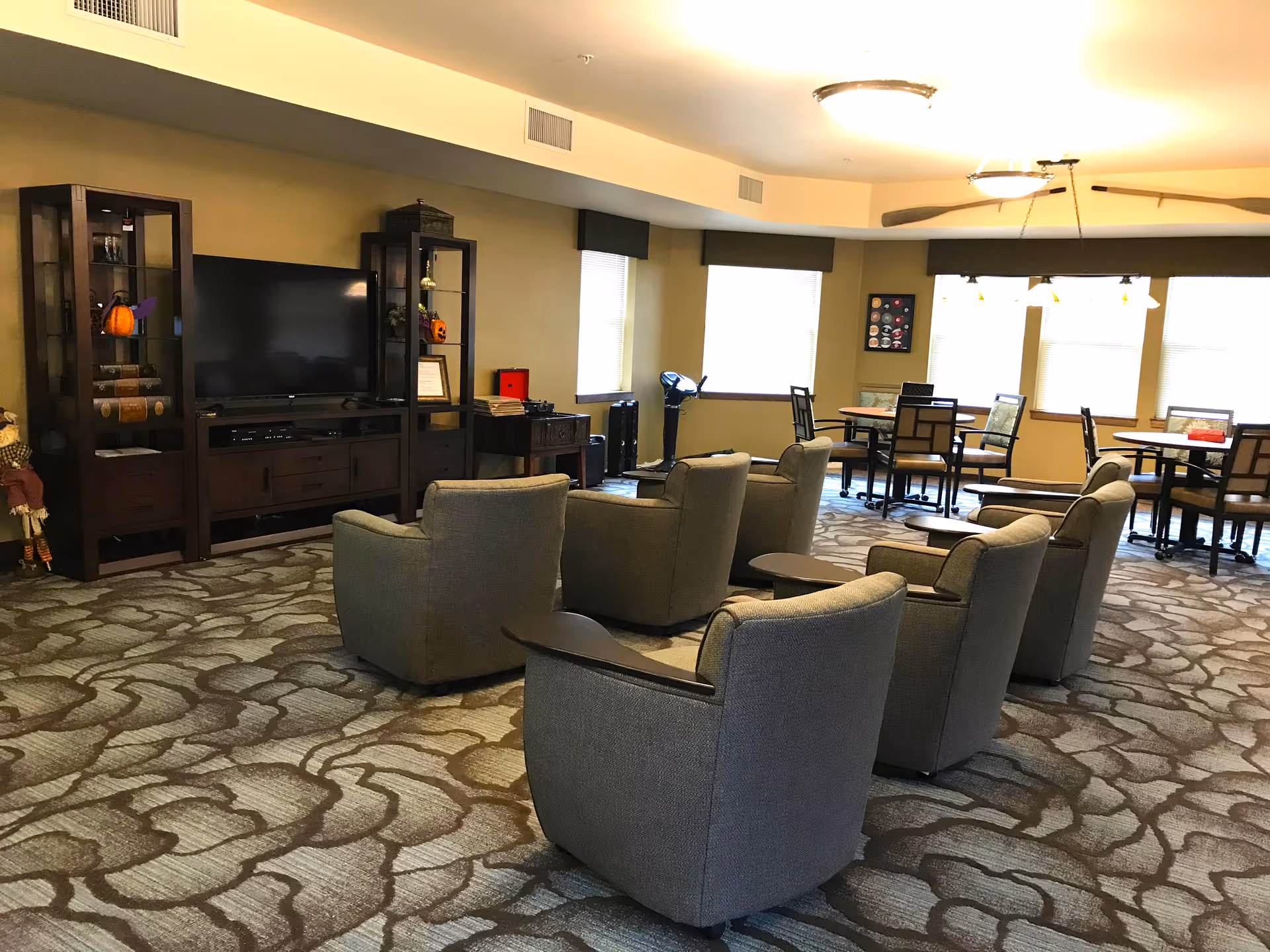 A senior living facility common area with several gray upholstered armchairs arranged facing a large flat-screen TV on a wooden entertainment center. The room has patterned carpet, multiple windows with blinds, and several tables with chairs in the background. The walls are painted beige and the ceiling has recessed lighting and a ceiling fan.