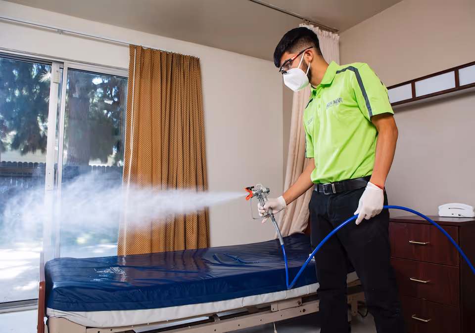A person wearing a green shirt, black pants, gloves, safety glasses, and a face mask is disinfecting a bed with a spray device in a room with a sliding glass door and brown curtains.
