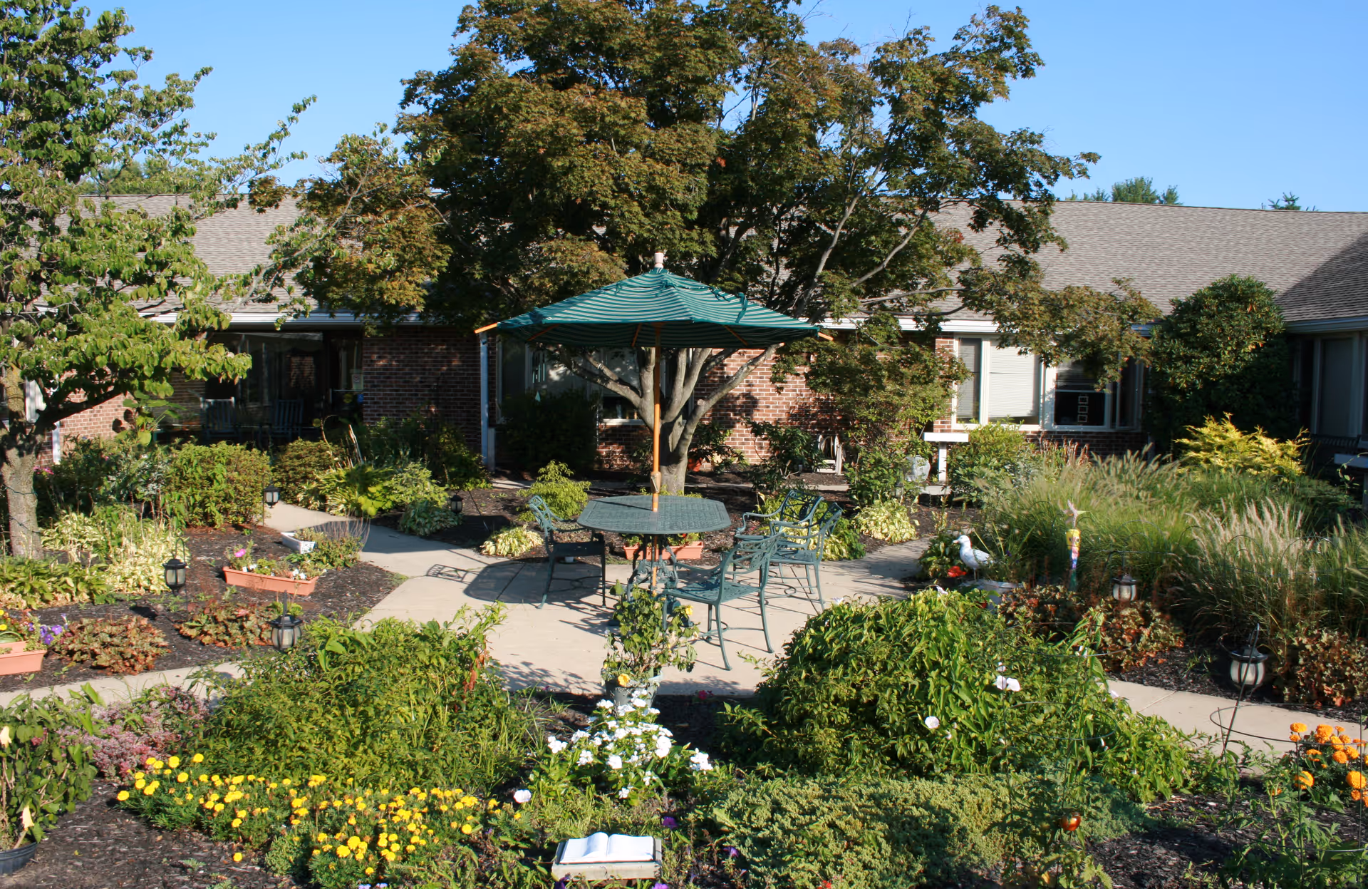 Outdoor garden area at Masonic Village at Warminster featuring a patio with a green umbrella over a round table surrounded by chairs, lush greenery, colorful flowers, and trees with a brick building in the background under a clear blue sky.