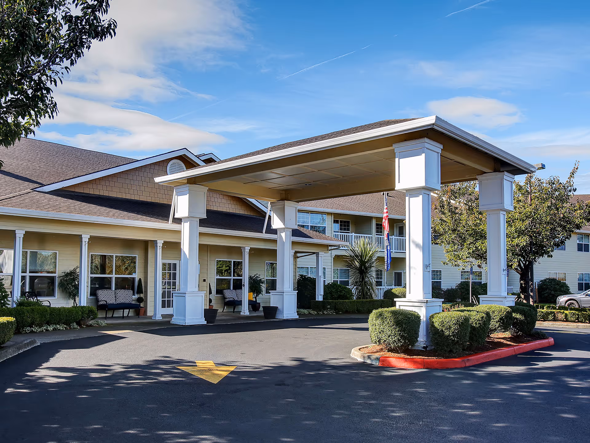 Exterior view of Prestige Senior Living Five Rivers showing the main entrance with a covered driveway supported by white pillars, surrounded by neatly trimmed bushes and trees under a blue sky with some clouds.