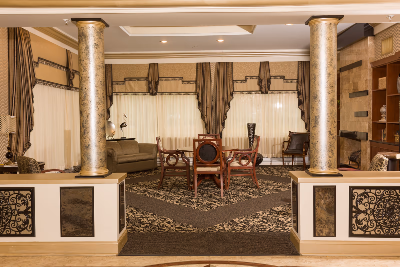 An elegant living room area with two large decorative columns in the foreground, a patterned carpet, a round wooden table with four chairs, a sofa, and additional chairs near large windows with sheer curtains and ornate drapes.