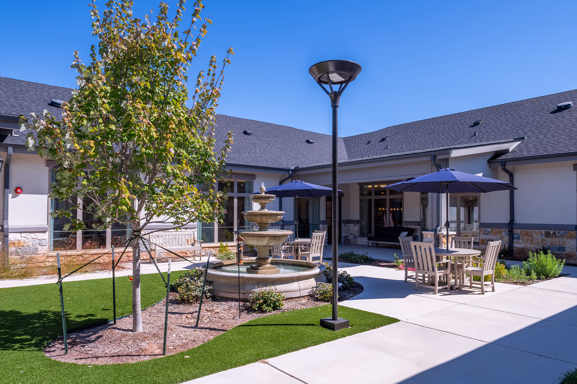 A sunny courtyard with a central tiered fountain, outdoor tables and umbrellas, a young tree and lamp post surrounded by the senior living building.