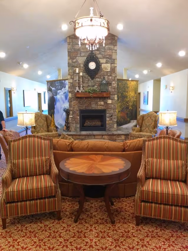 A cozy living room area in Turtle & Crane Assisted Living featuring a stone fireplace with a clock and decorative items on the mantel. In front of the fireplace are two patterned armchairs with striped upholstery, a round wooden table with a floral design, and a brown sofa. The room is well-lit with ceiling lights and two table lamps on either side of the fireplace. The carpet has a red and gold intricate pattern, and there are large nature-themed panels on either side of the fireplace.