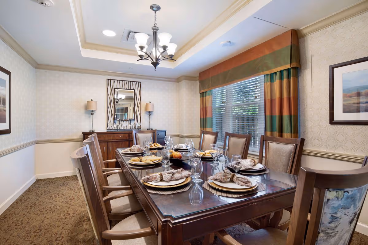 A formal dining room with a large wooden table set for eight people. The table is arranged with plates, napkins, glasses, and a centerpiece bowl with fruit. The room has patterned wallpaper, a window with striped curtains, a chandelier overhead, and a sideboard with a decorative mirror and two lamps.
