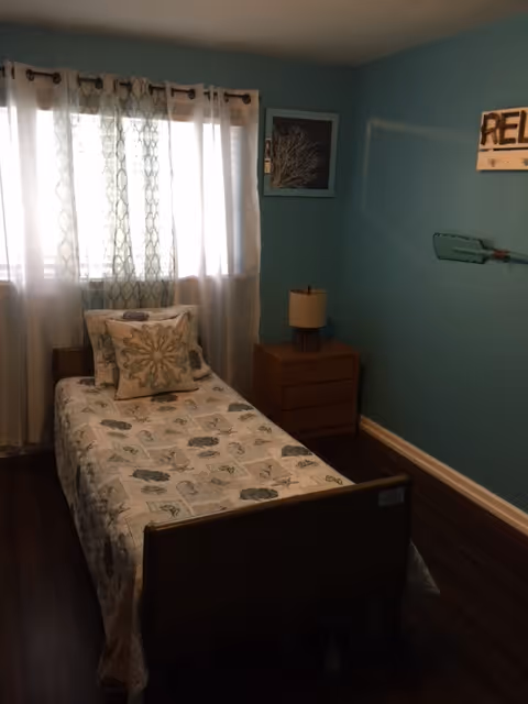 A small bedroom with a single bed covered in a patterned bedspread and decorative pillows. The room has teal walls, a wooden nightstand with a lamp, a window with sheer curtains, and wall decor including a framed picture and a sign that says 'RELAX'.