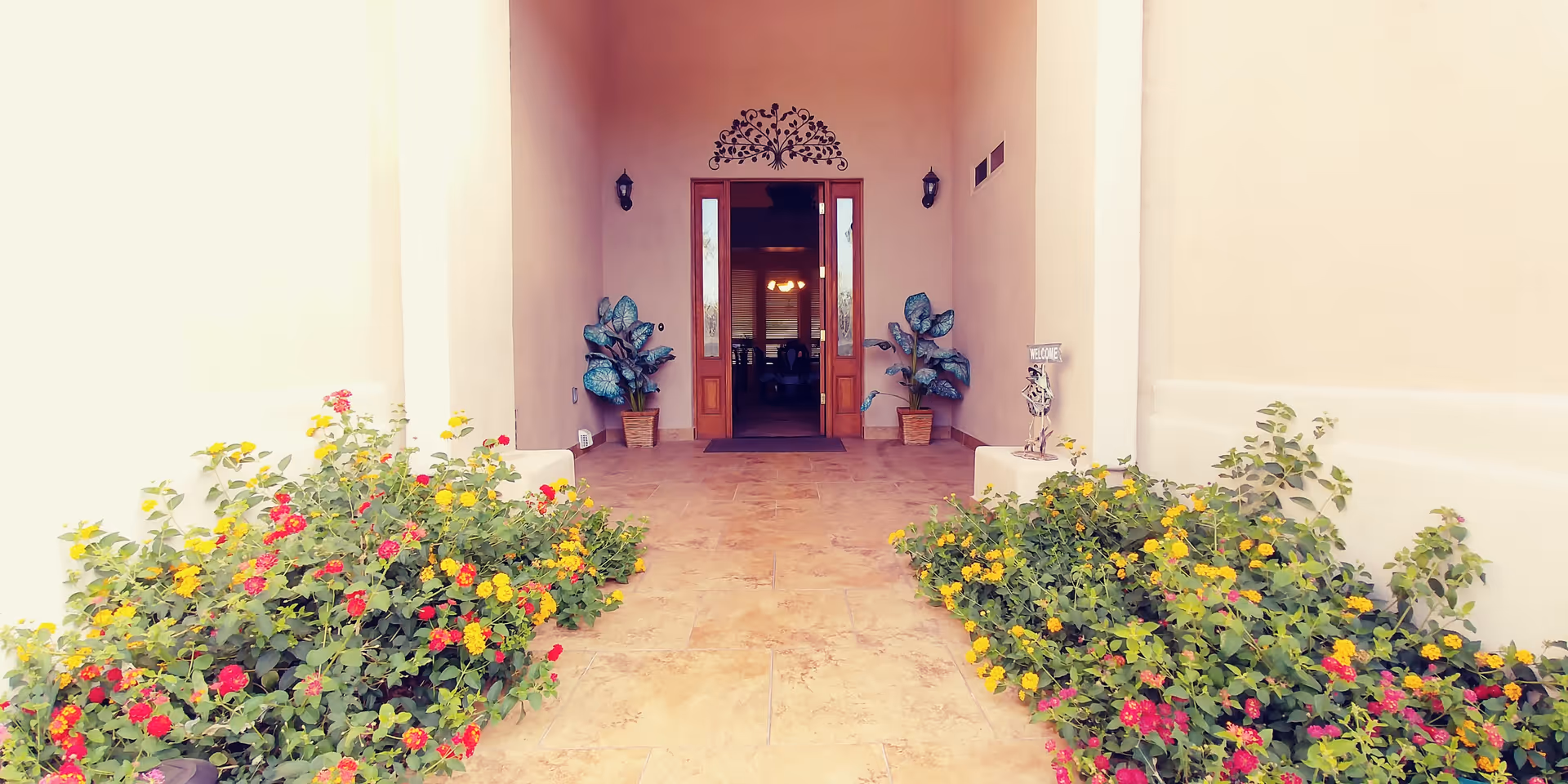 Entrance to a building with a tiled floor and beige walls, flanked by colorful flower bushes on both sides. Two potted plants are placed near the wooden double doors, which are open, revealing a glimpse of the interior with a chandelier and furniture.