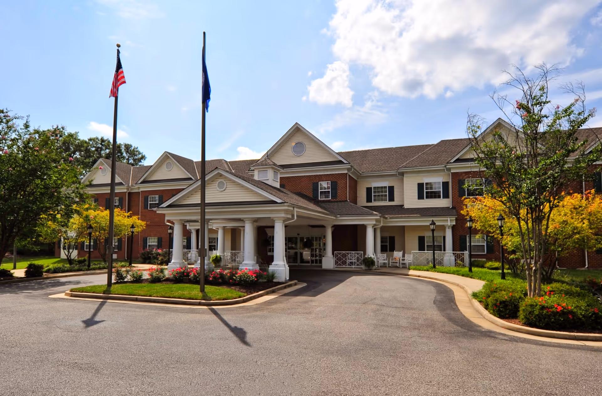 Front exterior view of a two-story senior living facility building with a covered entrance supported by white columns. There are two flagpoles with flags in front of the building, surrounded by landscaped greenery and flowers under a partly cloudy sky.