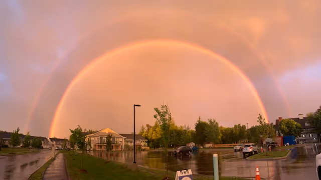 A vivid double rainbow arches across an orange sky over a wet parking lot and residential buildings.