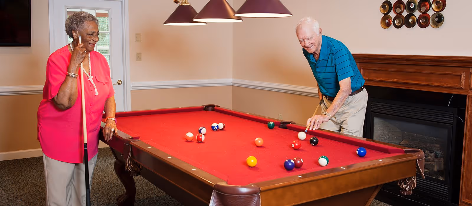 An elderly woman in a pink shirt and beige pants holding a pool cue and smiling while standing next to a pool table with a red felt surface. An elderly man in a blue striped polo shirt and beige pants is leaning over the table, aiming to hit a white cue ball. The room has beige walls, a fireplace with a wooden mantel, and a decorative circular wall piece above the fireplace.