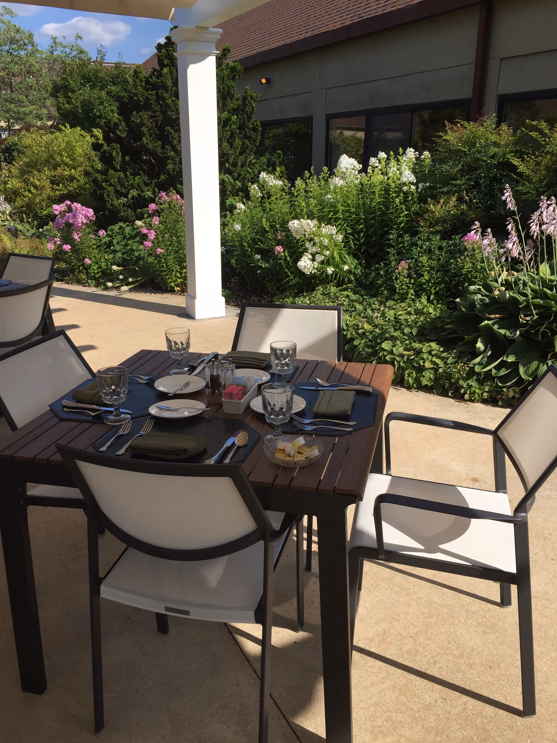 Outdoor patio dining area with a wooden table set for four, including plates, glasses, silverware, and napkins. The table is surrounded by four white and black chairs. In the background, there are green bushes and flowering plants next to a building with large windows under a clear blue sky.
