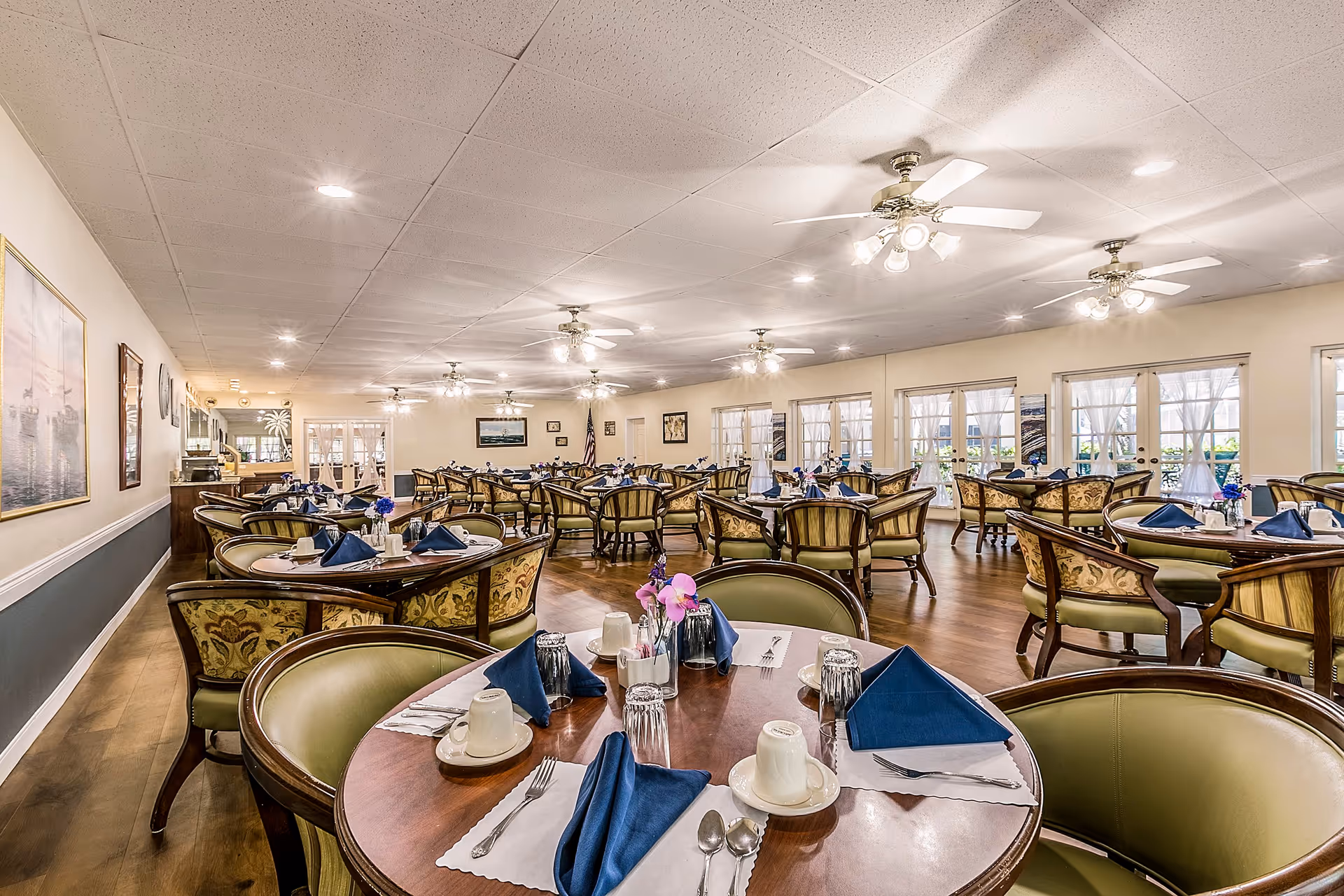 A spacious dining room with multiple round wooden tables set with white placemats, blue folded napkins, cups, glasses, and silverware. The room has green cushioned chairs with wooden frames, ceiling fans with lights, large windows with sheer white curtains, and framed artwork on the walls.