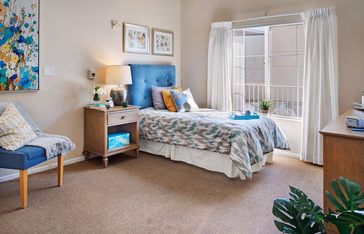 A bright and cozy bedroom with a single bed featuring a blue upholstered headboard and patterned bedding. Next to the bed is a wooden nightstand with a lamp, a small plant, and an alarm clock. A blue chair with a throw blanket and pillow is positioned to the left. The room has beige walls adorned with colorful floral artwork and two framed pictures above the bed. A large window with white curtains lets in natural light, and a wooden dresser is partially visible on the right side. A green plant is in the foreground.