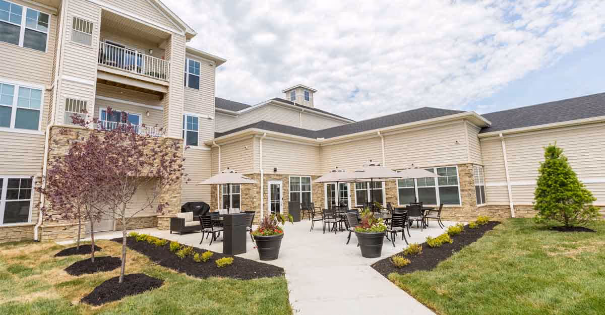 Outdoor patio with tables, umbrellas, planters and seating in front of a multi-story senior living building.