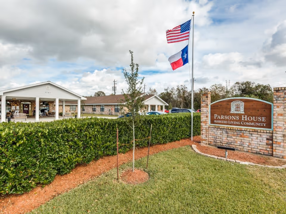 Exterior view of Parsons House Assisted Living Community showing a brick sign with the facility name, two flagpoles with American and Texas flags, a green hedge, a small tree, and a building with a covered entrance and rocking chairs under a cloudy sky.